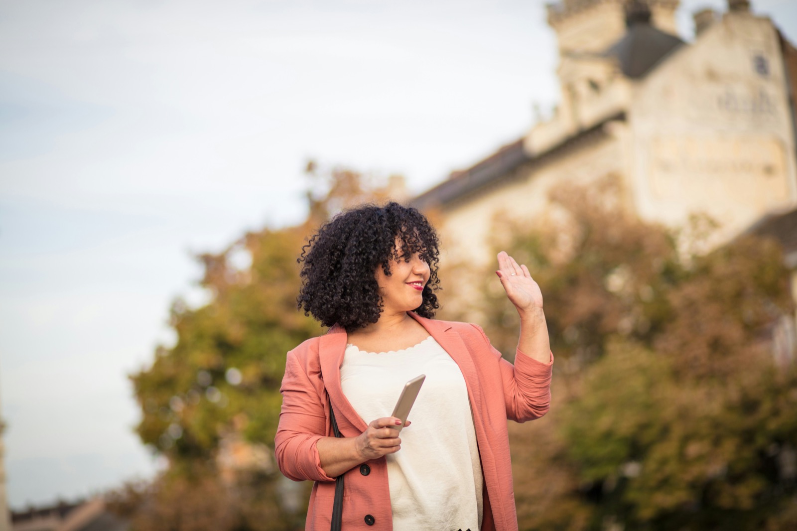 A woman holding a phone waves hello outdoors on an autumn day.
